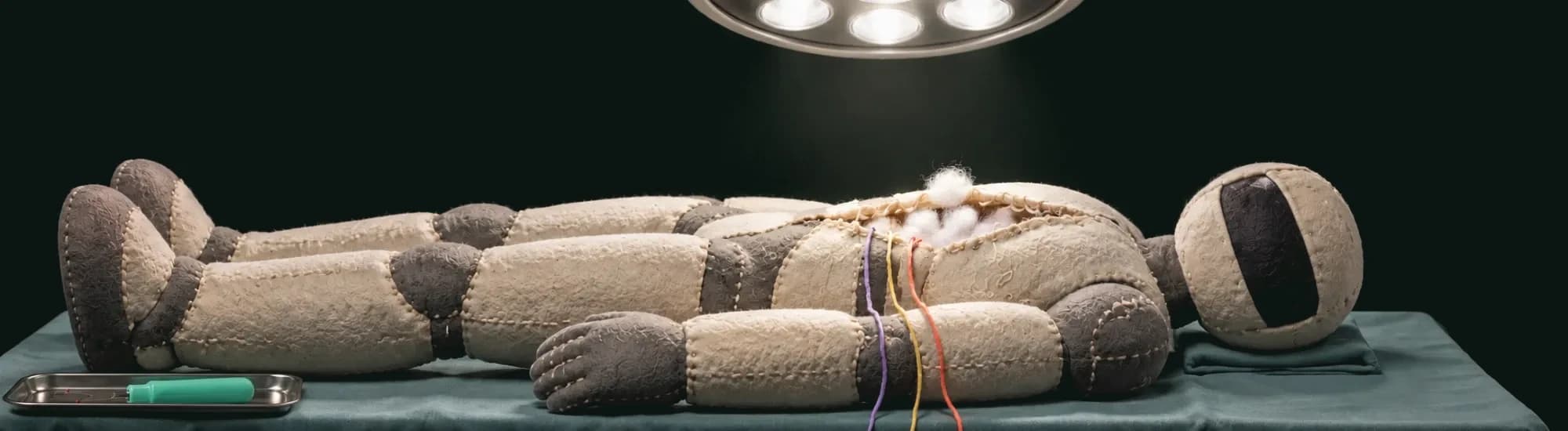 A hand-sewn felt robot lies on an operating table under a surgical lamp. Its chest seam is unpicked and the stuffing shows.